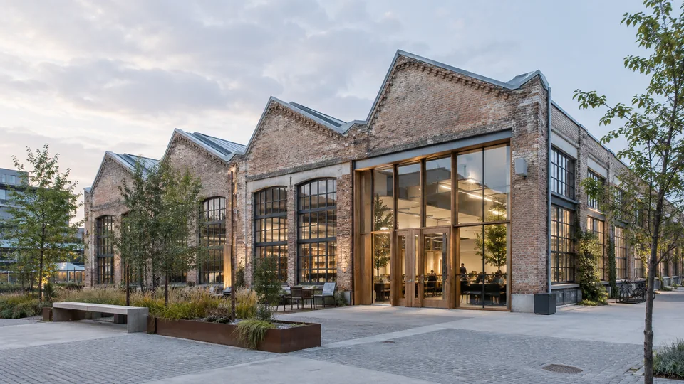 Illustration d’une ancienne halle industrielle en brique reconvertie, avec toiture en sheds et nouveaux espaces vitrés.